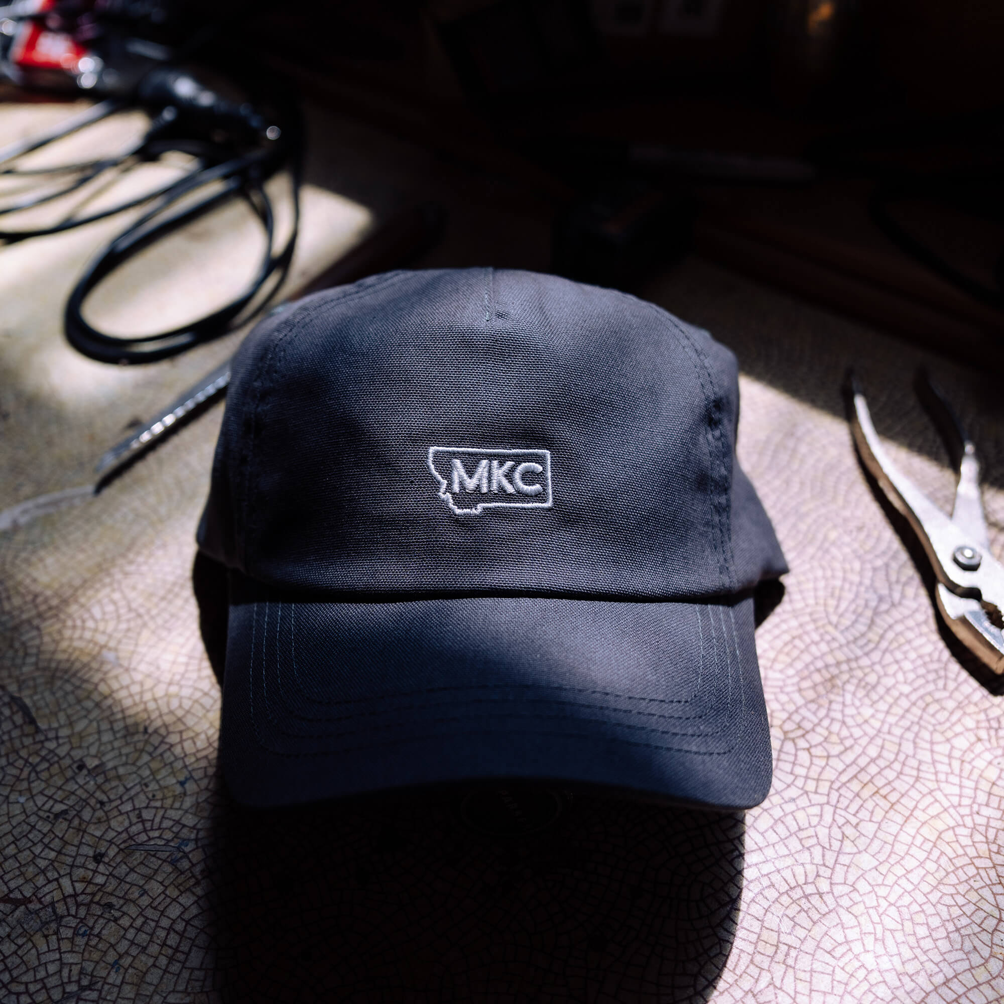 Grey MKC canvas dad hat featuring embroidered logo, placed on a textured surface with tools in the background.