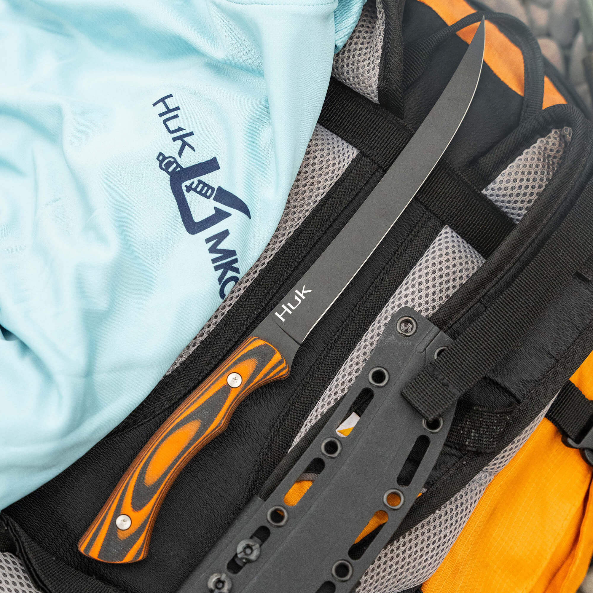HUK Flathead fillet knife with a black blade and orange handle resting on a backpack and a light blue shirt.
