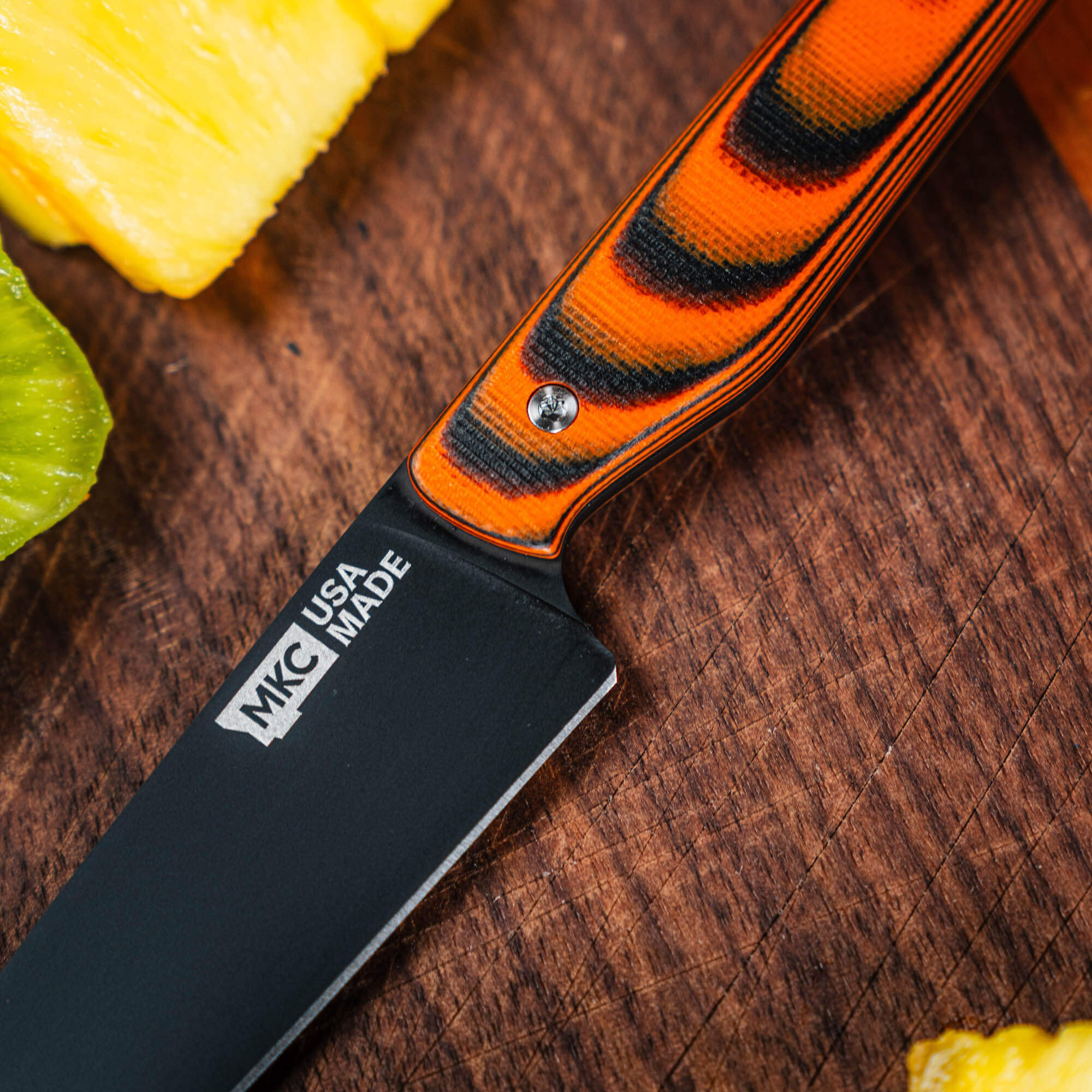 Close-up of the CUTBANK paring knife with an orange and black handle, resting on a wooden cutting board with fresh produce.