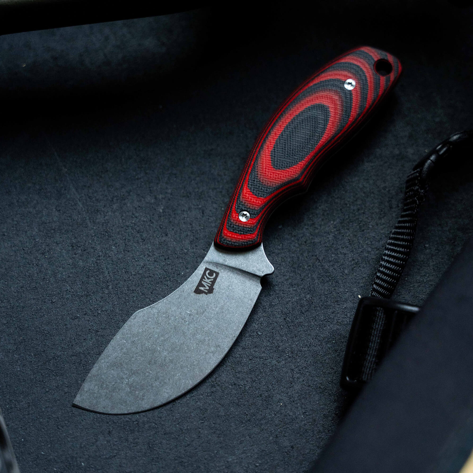 Alt text: Close-up of a Packout Skinner knife with a red and black handle and a matte grey blade, marked with the logo "MKC", displayed on a dark background.