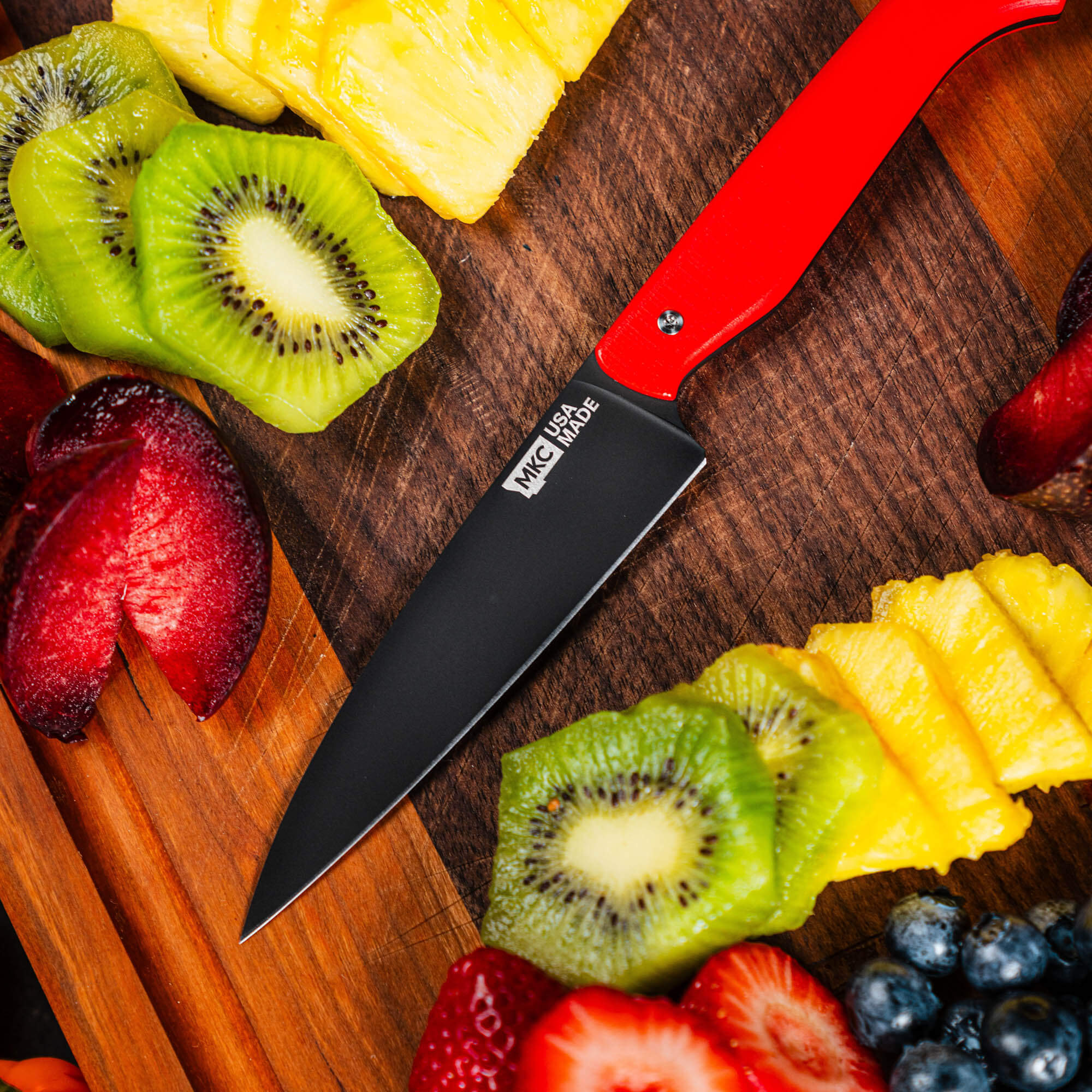 Red CUTBANK paring knife on a wooden cutting board surrounded by fresh fruits like kiwi, pineapple, strawberries, and blueberries.