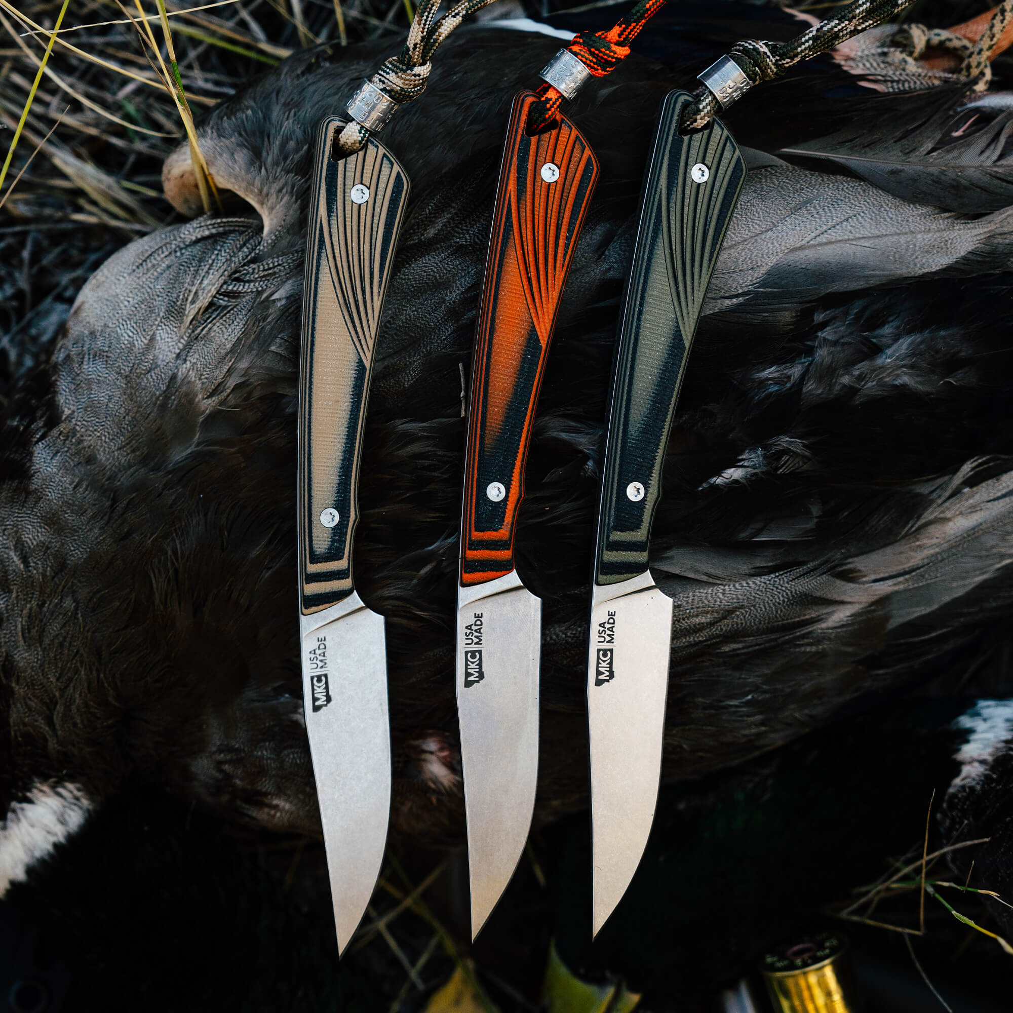Three CHÊNE Freezout knives featuring unique handle designs in orange, green, and brown, resting on dark feathers.
