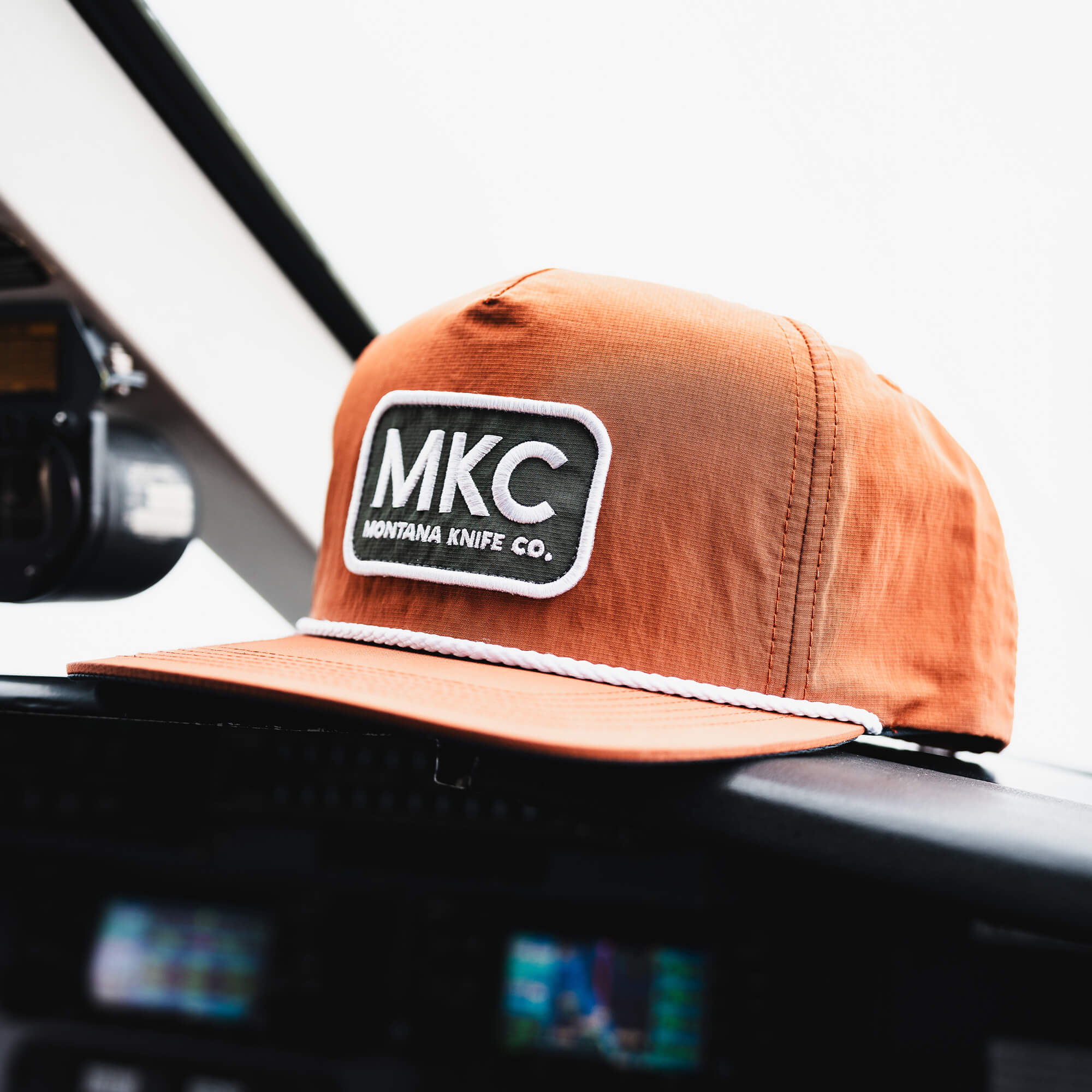 Rust-colored MKC River Rope Hat featuring a logo patch, set on a cockpit dashboard. Ideal for outdoor enthusiasts.