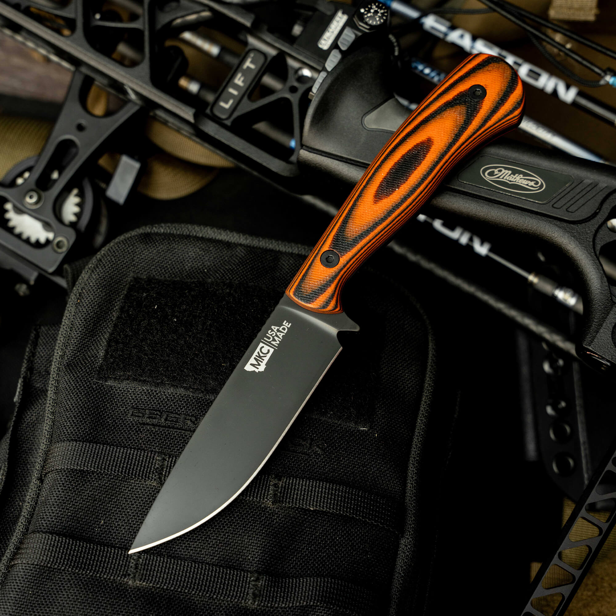 Mathews Archery MKC Whitetail knife with orange and black handle, placed on a tactical pouch and bow backdrop.
