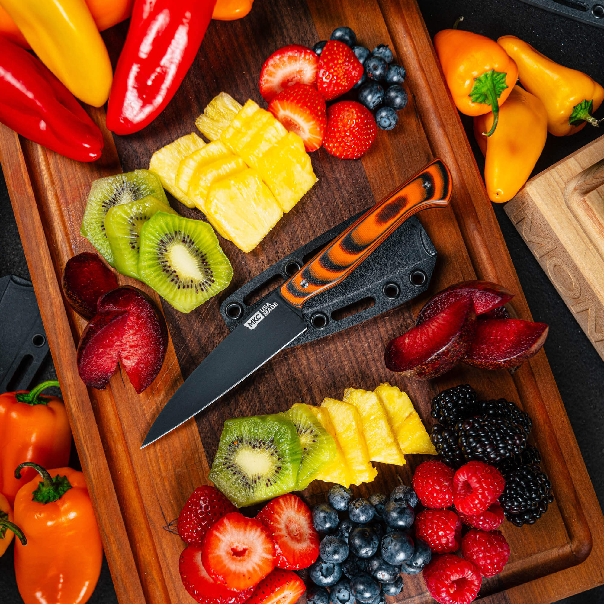 Montana Knife Company's Cutbank Paring Knife in orange and black, displayed with a variety of fresh fruits on a wooden cutting board.