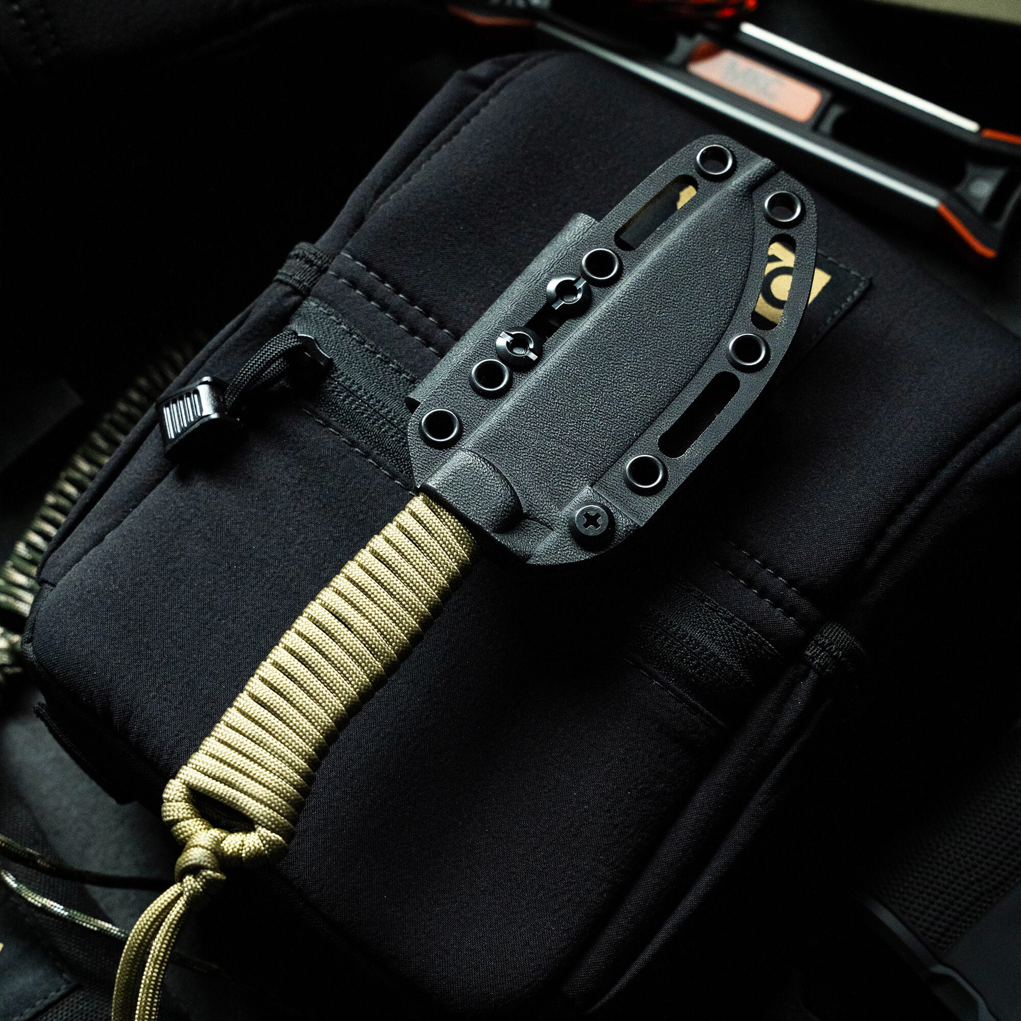 Black tactical knife secured in a sheath, attached to a black bag with gold and black detailing, part of the Wild Sheep Foundation Stoned Goat 2.0 collection.