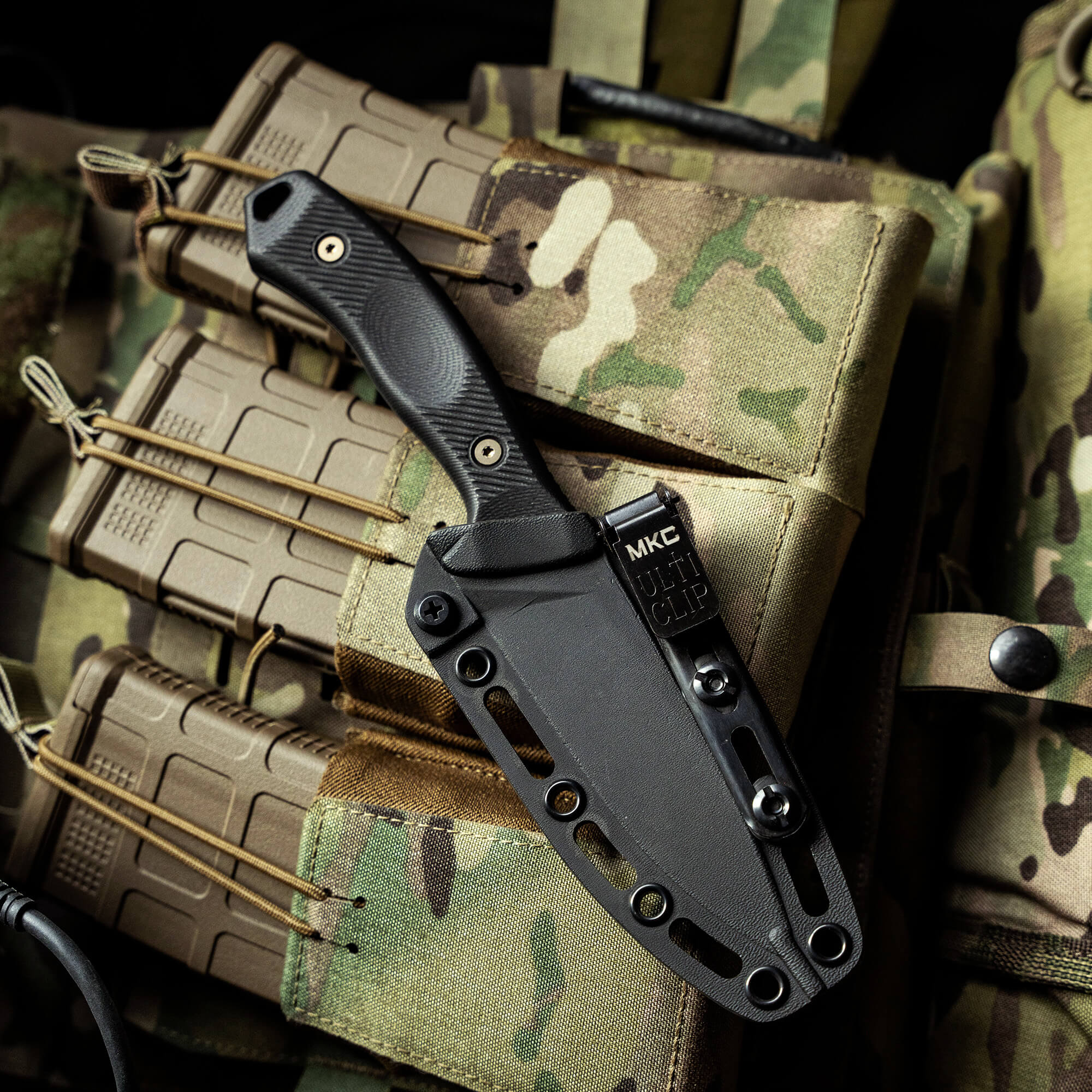 Black Kydex sheath with a partially visible knife handle attached to a camouflage tactical vest, highlighting the sheath's durability and secure attachment features.