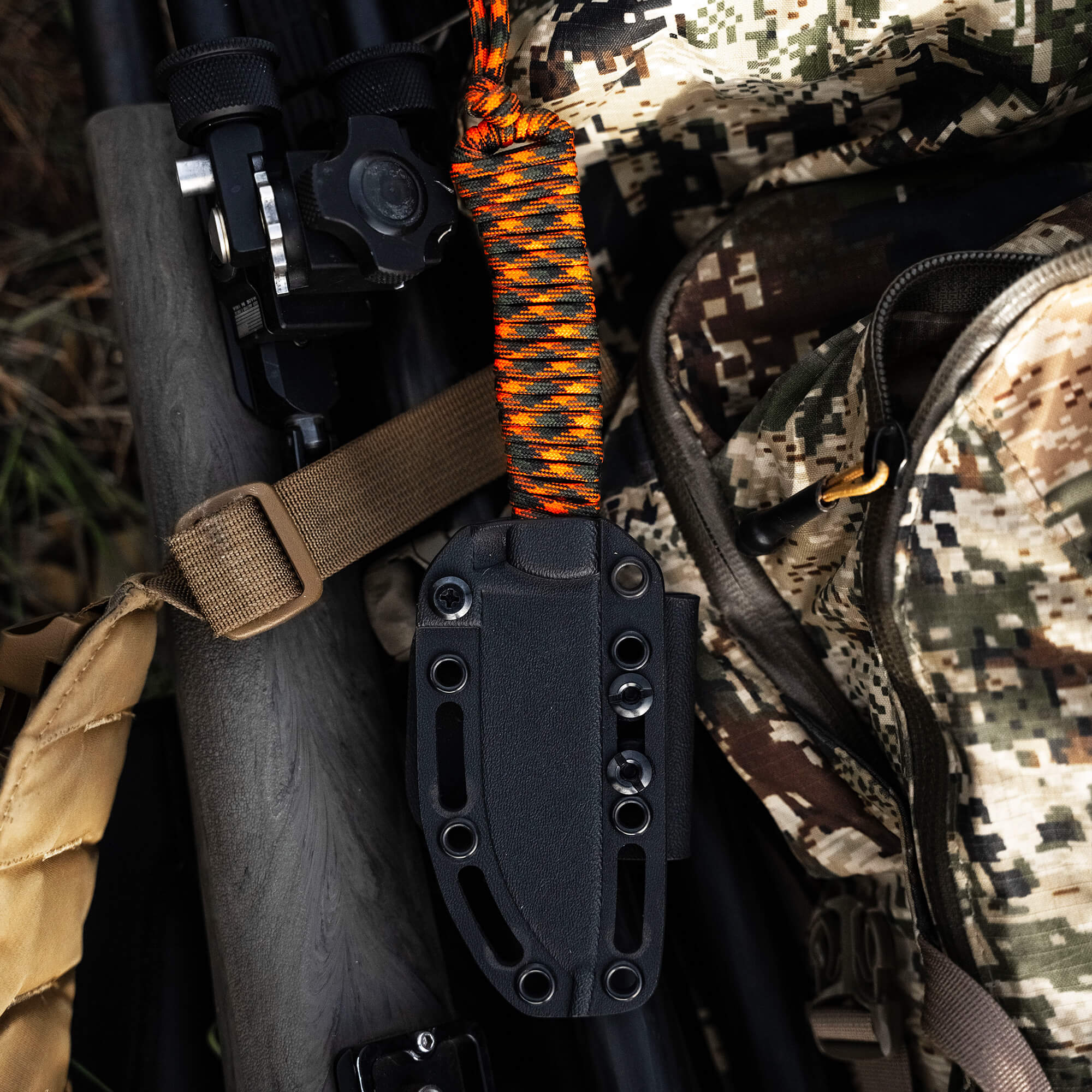 A close-up view of a Stoned Goat 2.0 knife in a black sheath attached to a tripod, with a camouflaged background featuring a backpack, binoculars, and multicolored paracord.