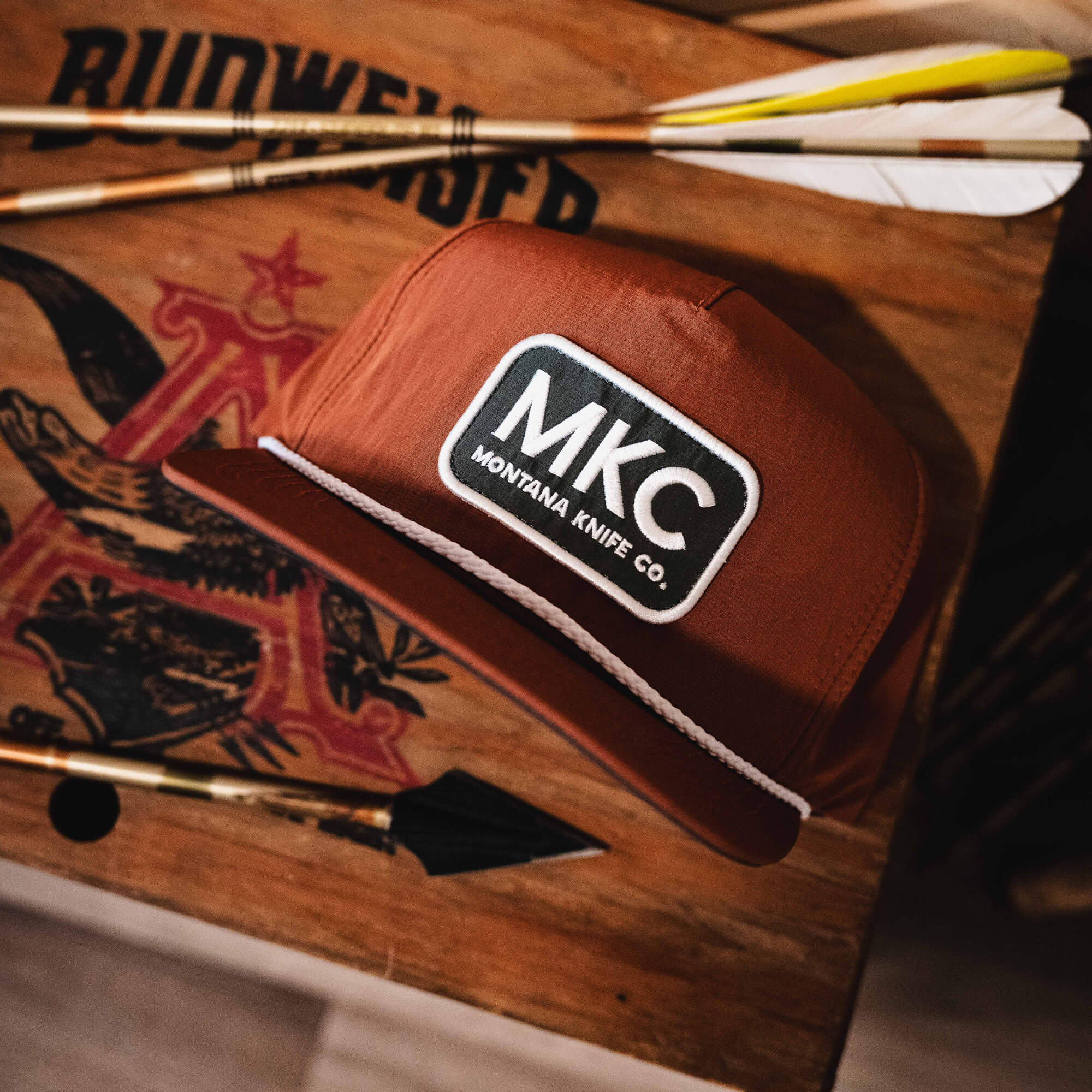 Rust-colored MKC River Rope Hat featuring a logo patch, placed on a wooden surface with arrows and tools.
