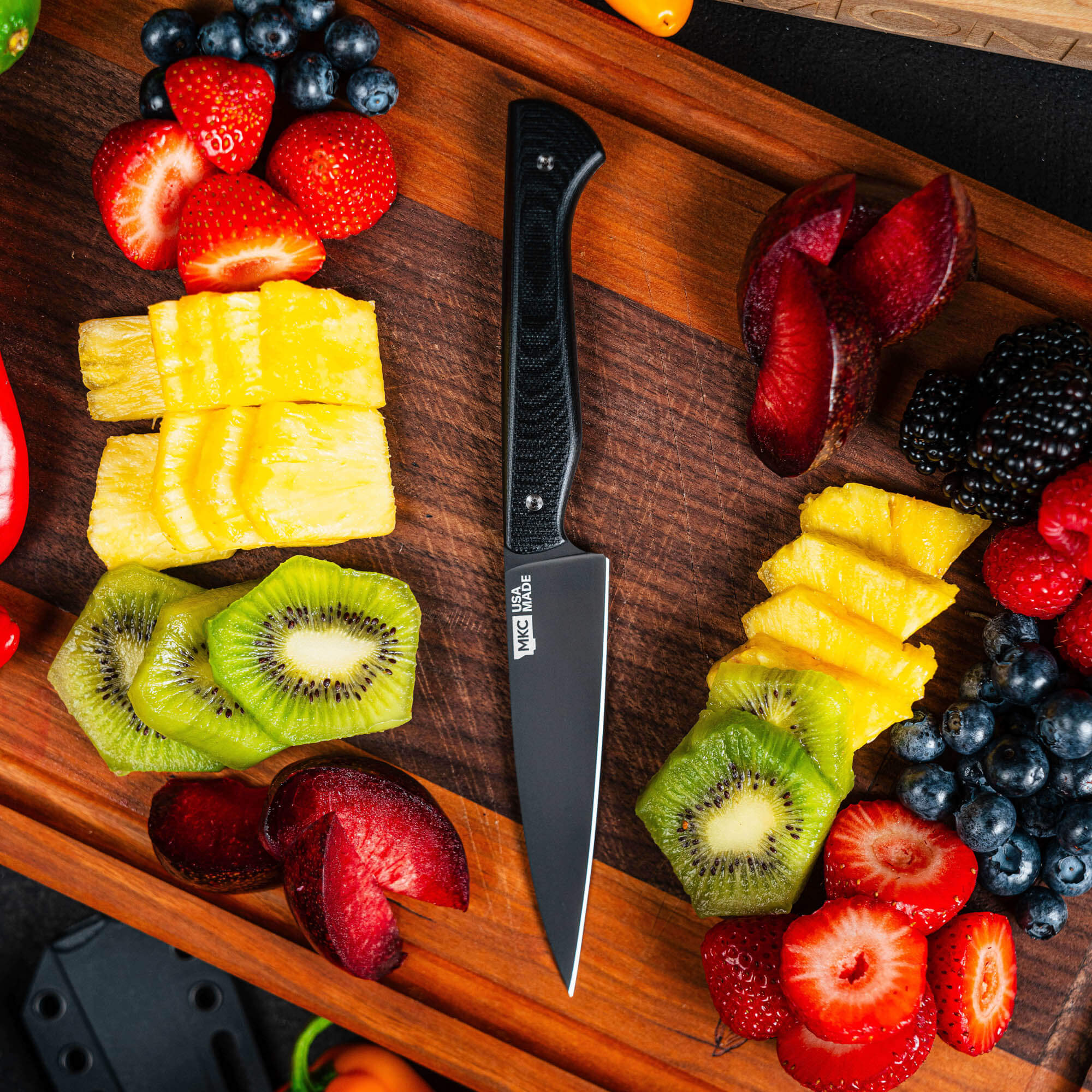 Black Cutbank paring knife on a wooden board surrounded by colorful sliced fruits including kiwi, pineapple, and berries.