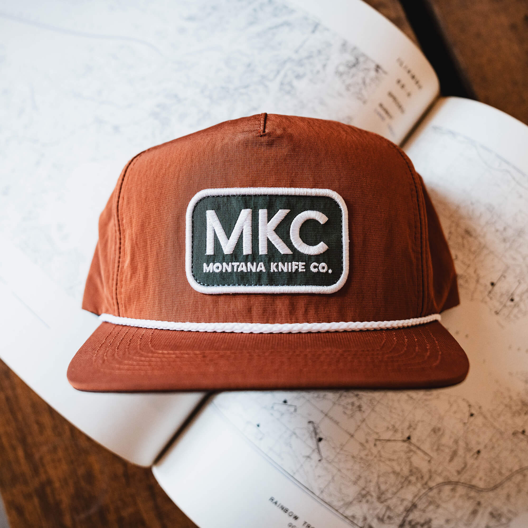 Rust-colored MKC River Rope Hat featuring a logo patch with Montana Knife Co branding, set on a map background.