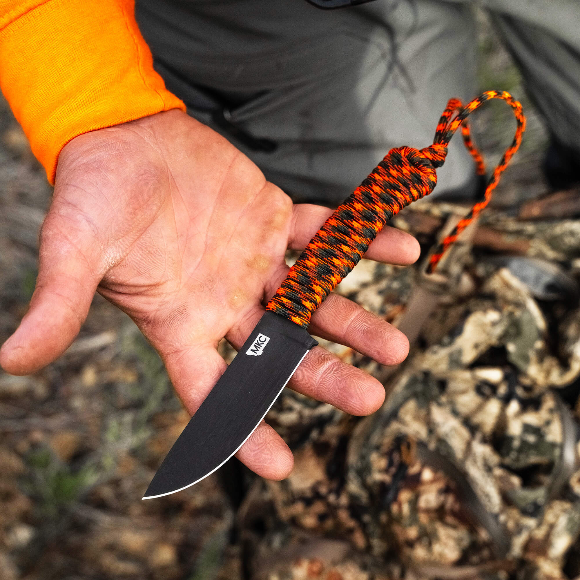 Alt text: "Close-up view of a hand holding the Wild Sheep Foundation Stoned Goat 2.0 knife, featuring a vibrant orange and black paracord-wrapped handle and a sleek, matte black blade, in an outdoor setting with blurred