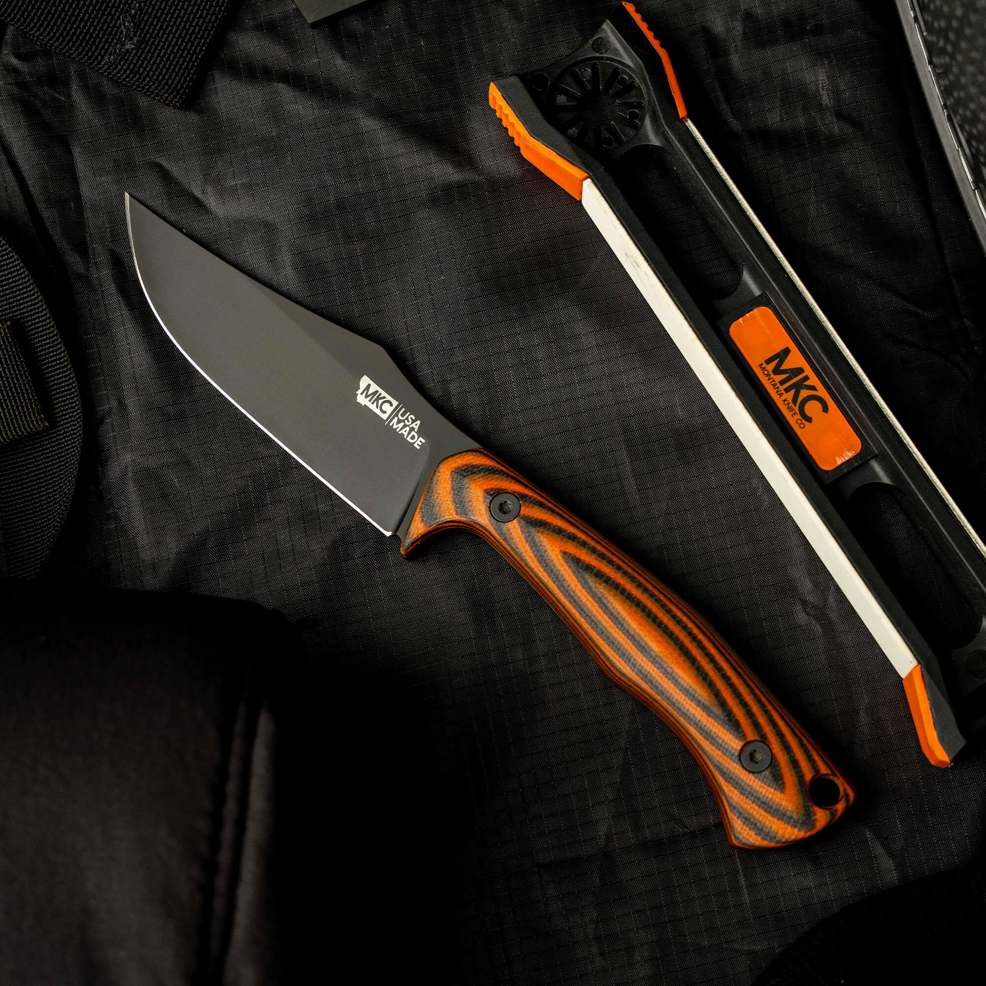Montana Knife Company's Nock On Triumph Pro knife with a black blade and orange striped handle, displayed on a textured surface.