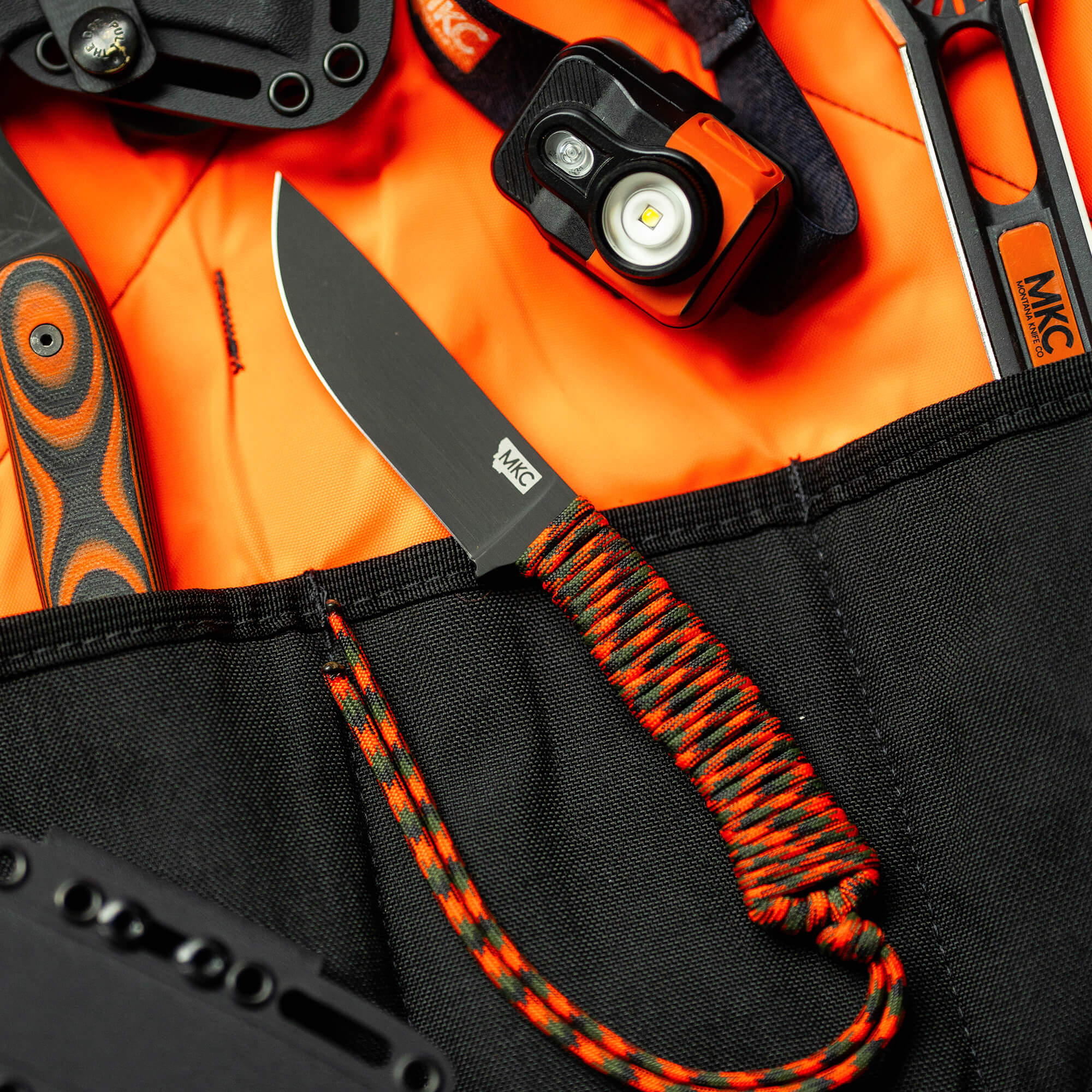 Alt text: Close-up view of a high-quality hunting knife with an orange and black handle, set against a bright orange fabric background. The image includes outdoor gear like a headlamp, another knife, and a black storage pouch, all showcasing the rugged