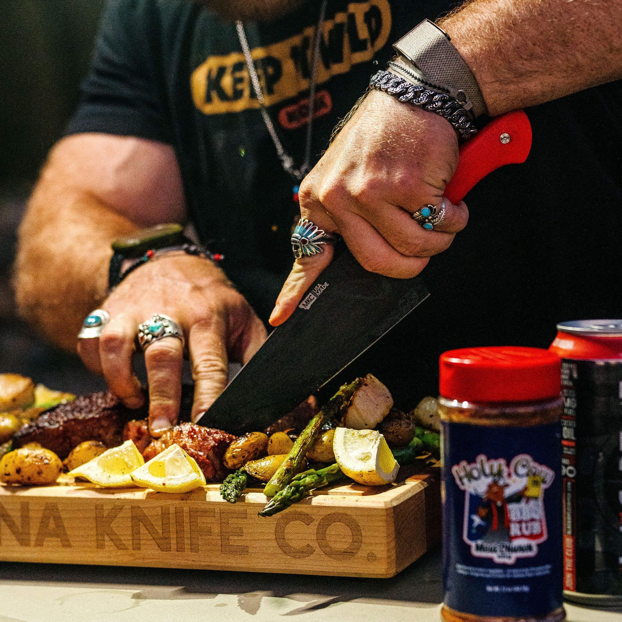Person slicing grilled meats and vegetables on a wooden board using a Bighorn Chef knife with a distinctive red handle, surrounded by seasonings and garnishes.