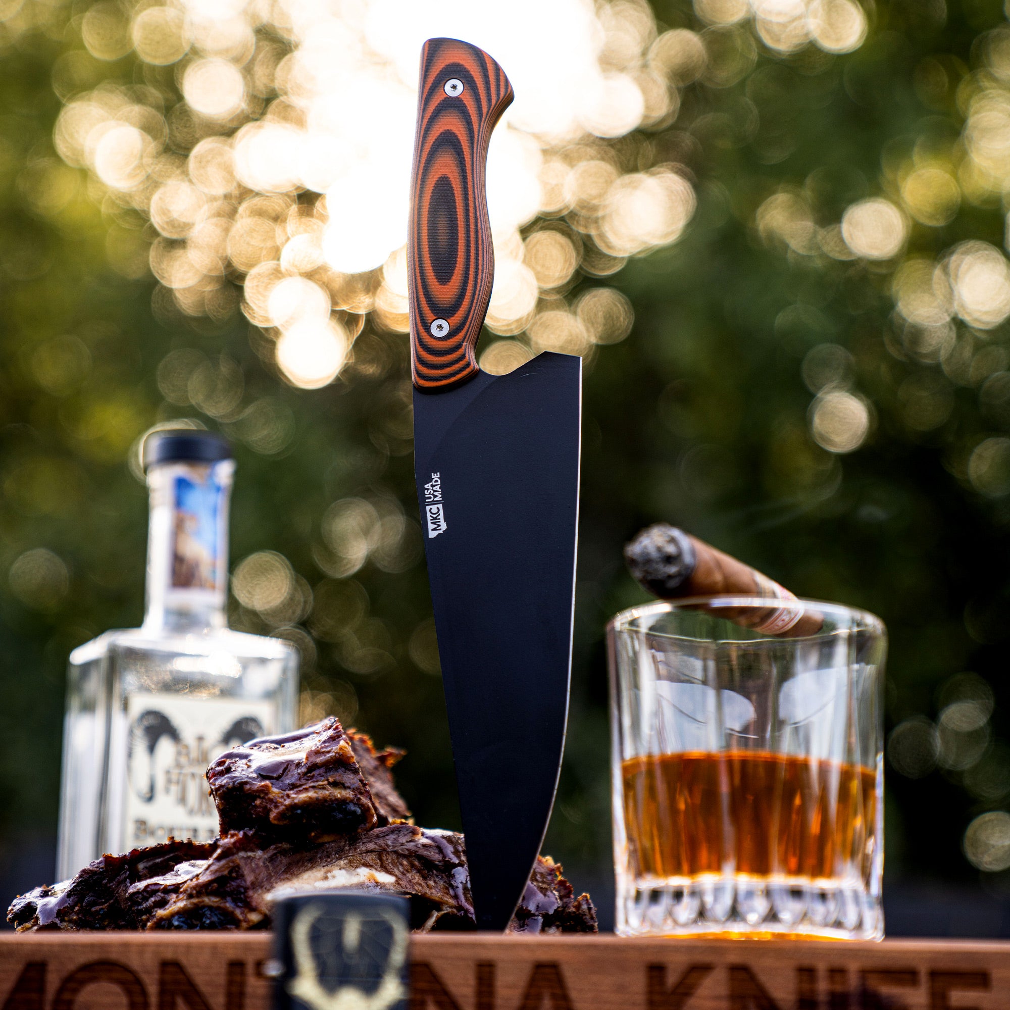 Alt text: A Bighorn Chef knife with an orange and black striped handle stands vertically on a wooden board, beside BBQ ribs, a glass of whiskey, and a cigar. The knife features a black blade with the Montana Knife logo etched near