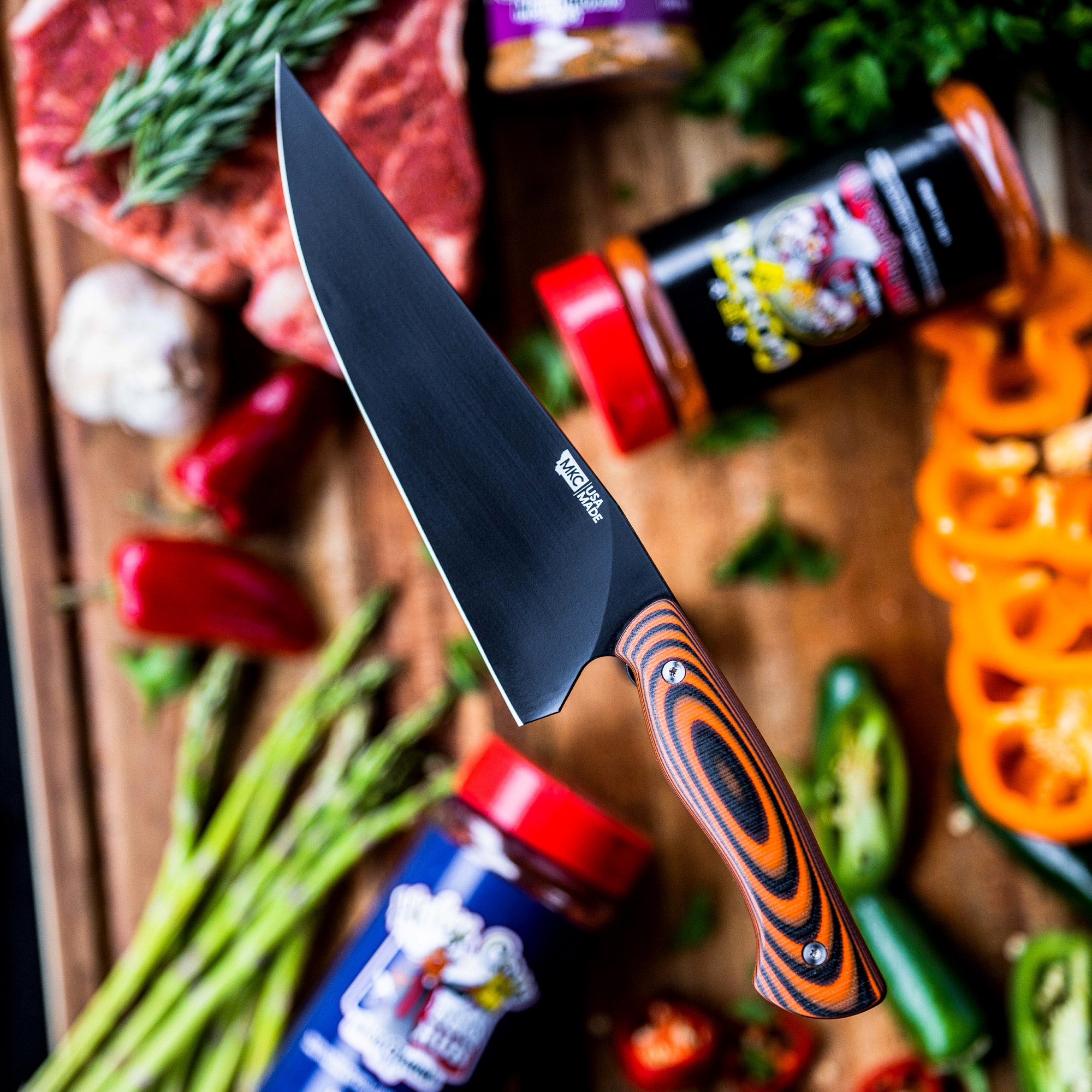 Alt text: "Close-up of a BIGHORN CHEF knife with an orange and black handle, featuring a sleek black blade, displayed on a wooden board surrounded by fresh vegetables and jars of spices."