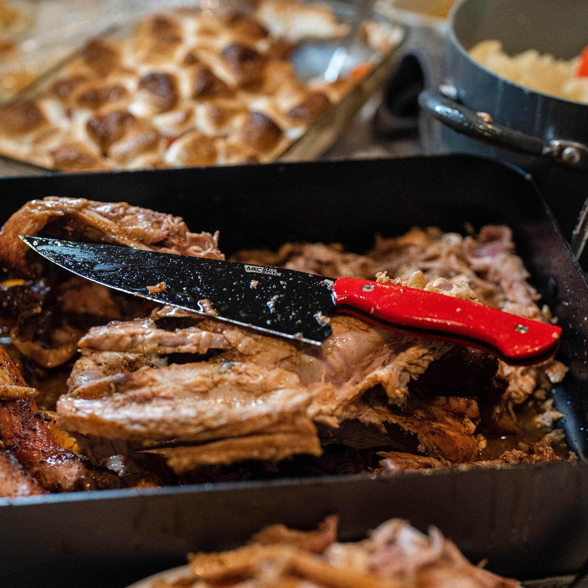 Red-handled Bighorn Chef knife resting on shredded pork in a cooking pan, surrounded by other cooking utensils and dishes.
