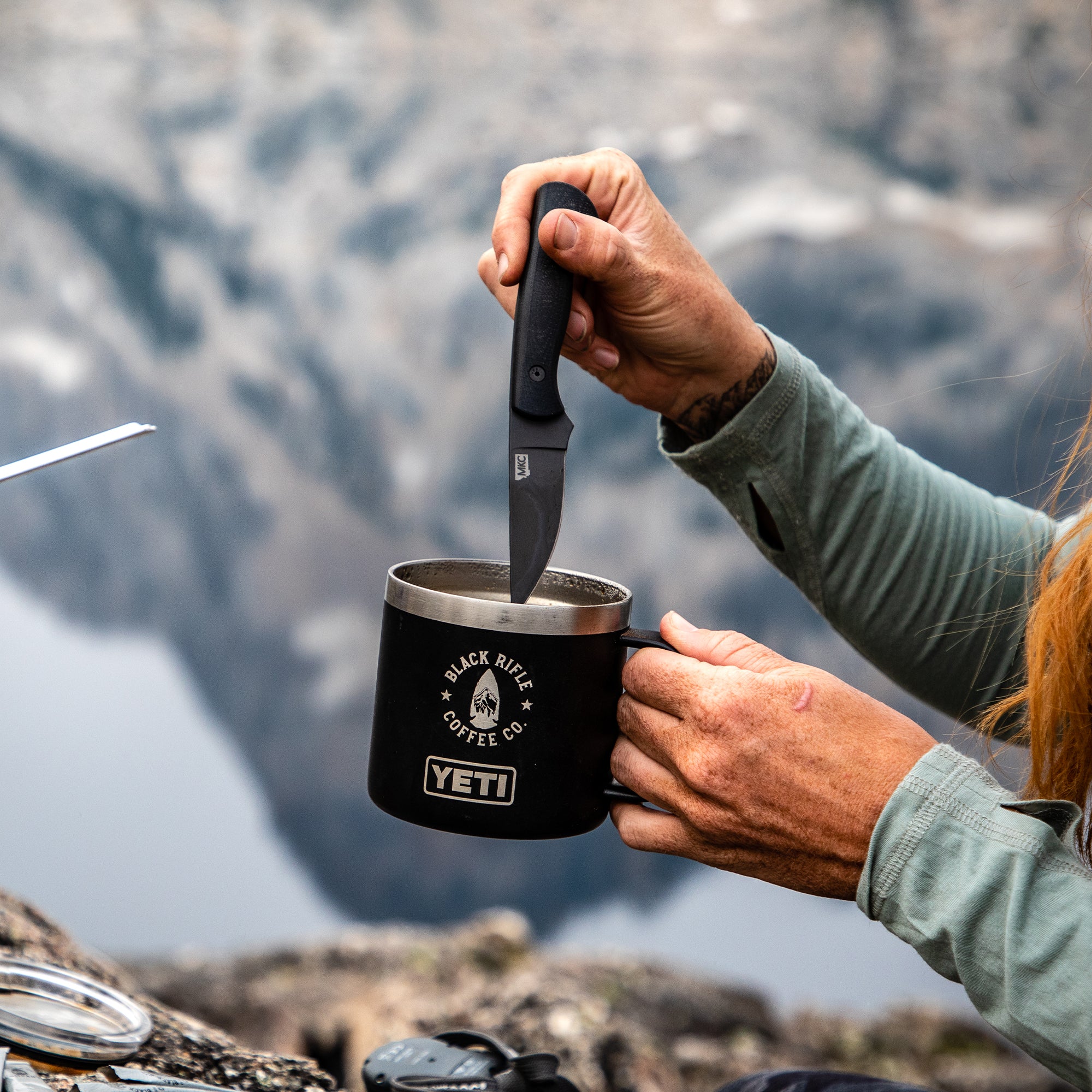 Alt text: Person using a BLACKFOOT 2.0 black tactical knife to stir a drink in a black YETI mug adorned with Black Rifle Coffee Co. logo, with a rugged mountain landscape in the background.