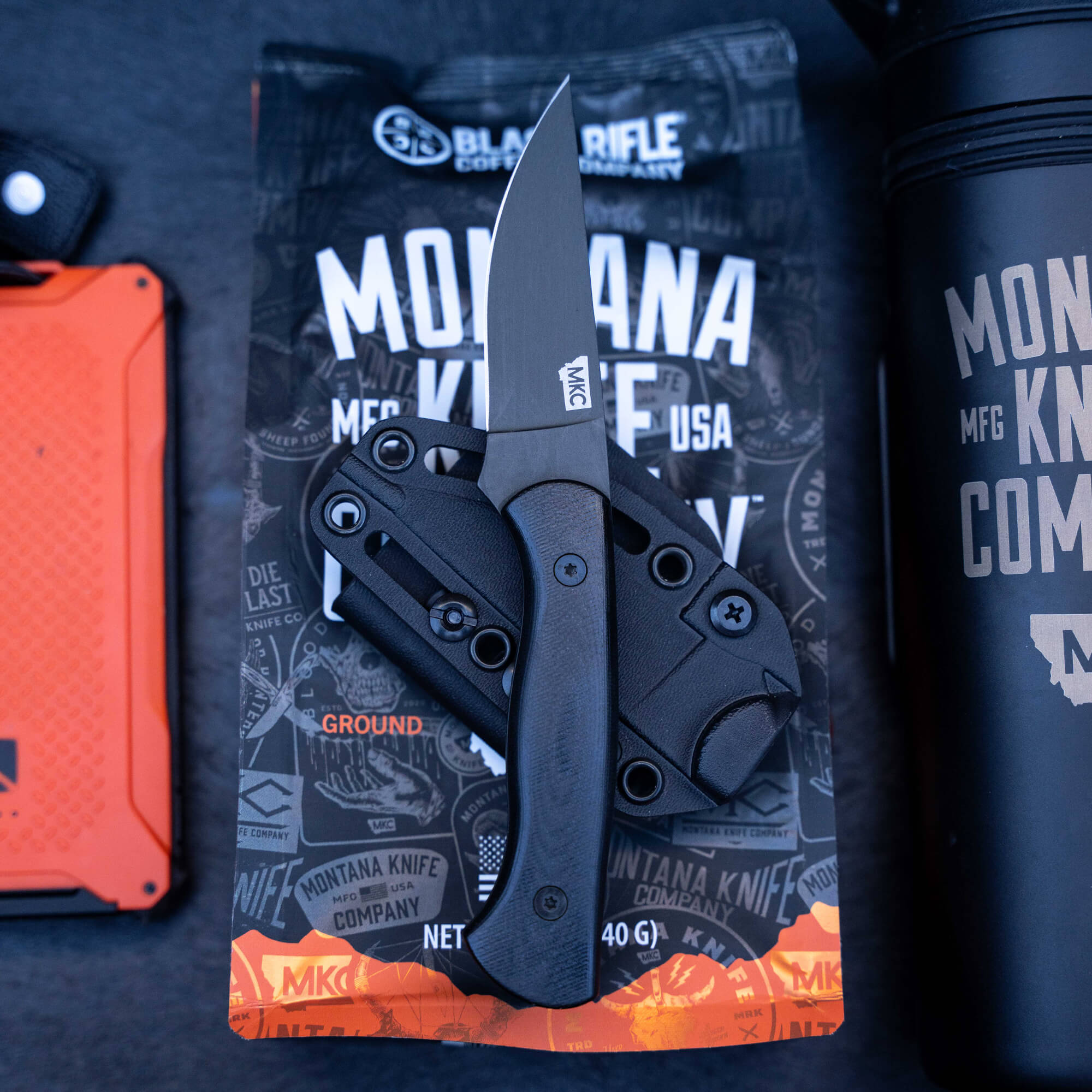 Alt text: "Blackfoot 2.0 tactical knife by Montana Knife Company, displayed open on a branded packaging with logos, accompanied by outdoor gear including an orange case and black thermal bottle, emphasizing durability and American manufacturing."