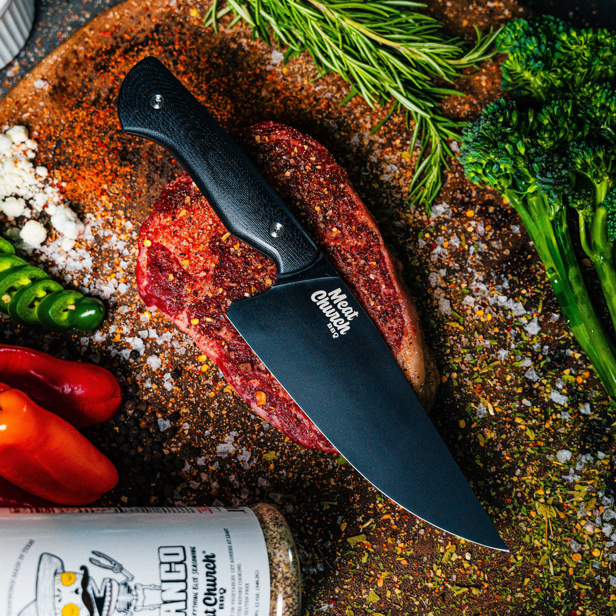 Meat Church chef knife on a cutting board with steak, spices, and fresh vegetables, showcasing its sharp blade and ergonomic handle.