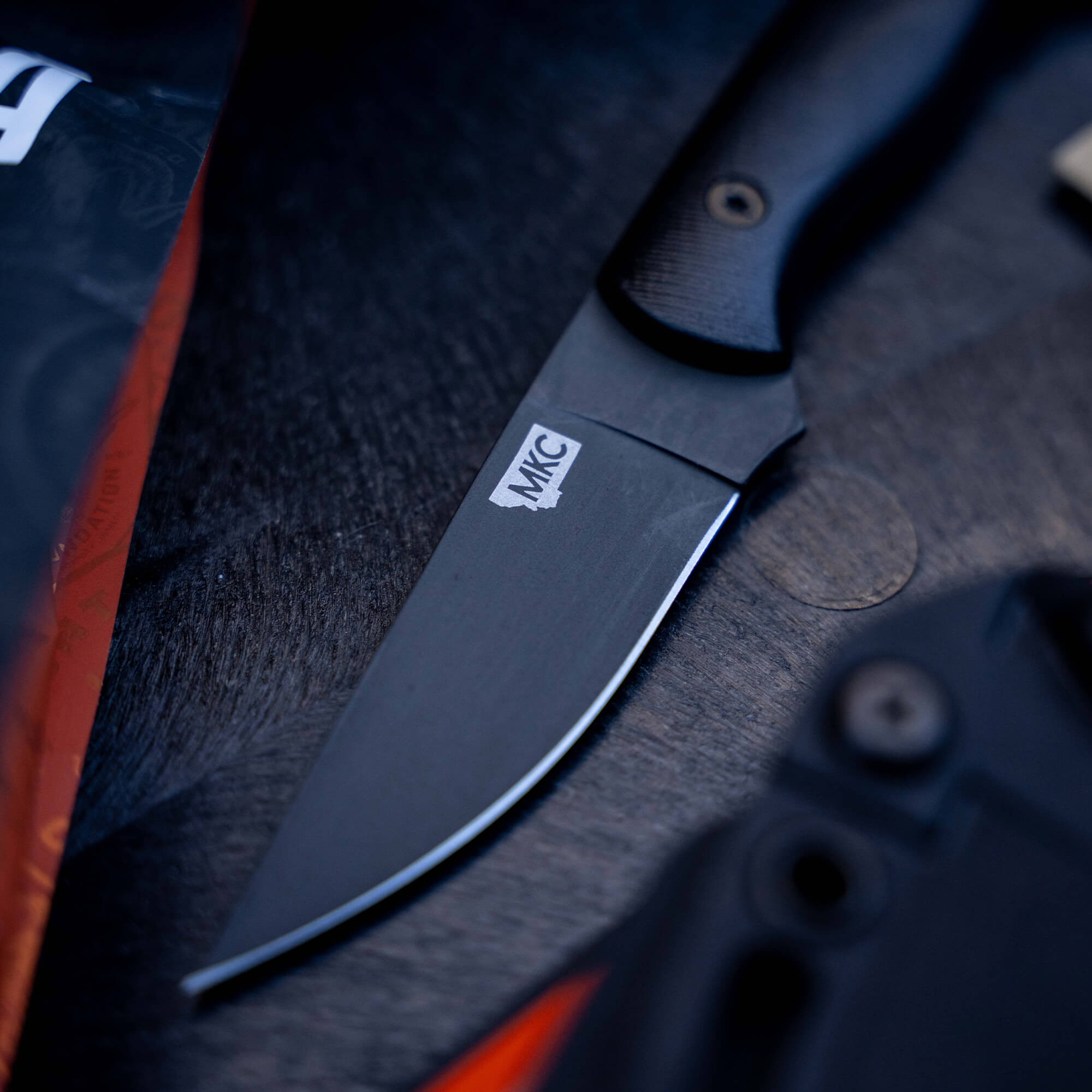 Close-up image of a BLACKFOOT 2.0 tactical knife in black color, featuring a matte blade with the MKC logo on it, positioned on a textured dark surface with soft-focus background elements.