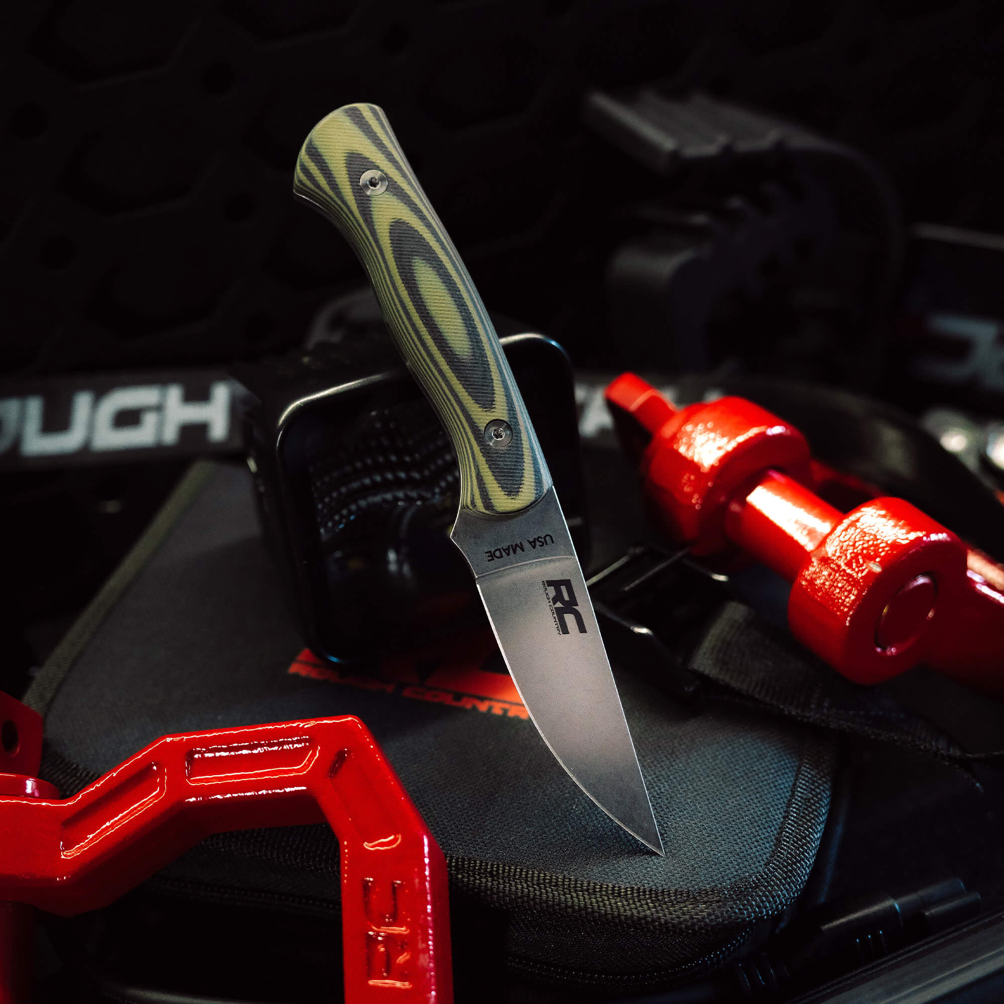 Rough Country Magnacut Blackfoot 2.0 knife with a patterned handle, placed on a gear backdrop with weights.