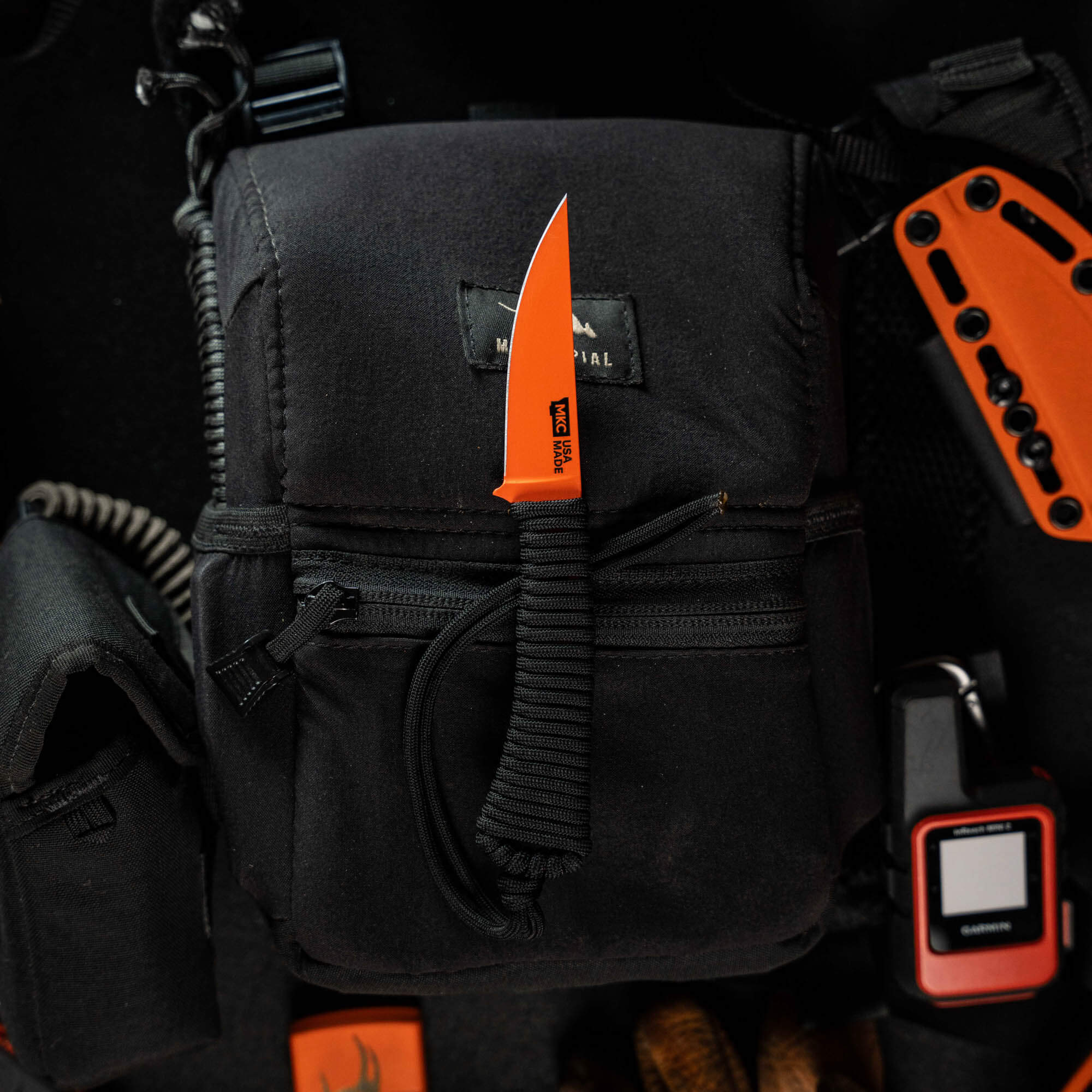 Mini Speedgoat 2.0 knife with Blaze Orange Cerakote, resting on a black bag among outdoor gear and tools.