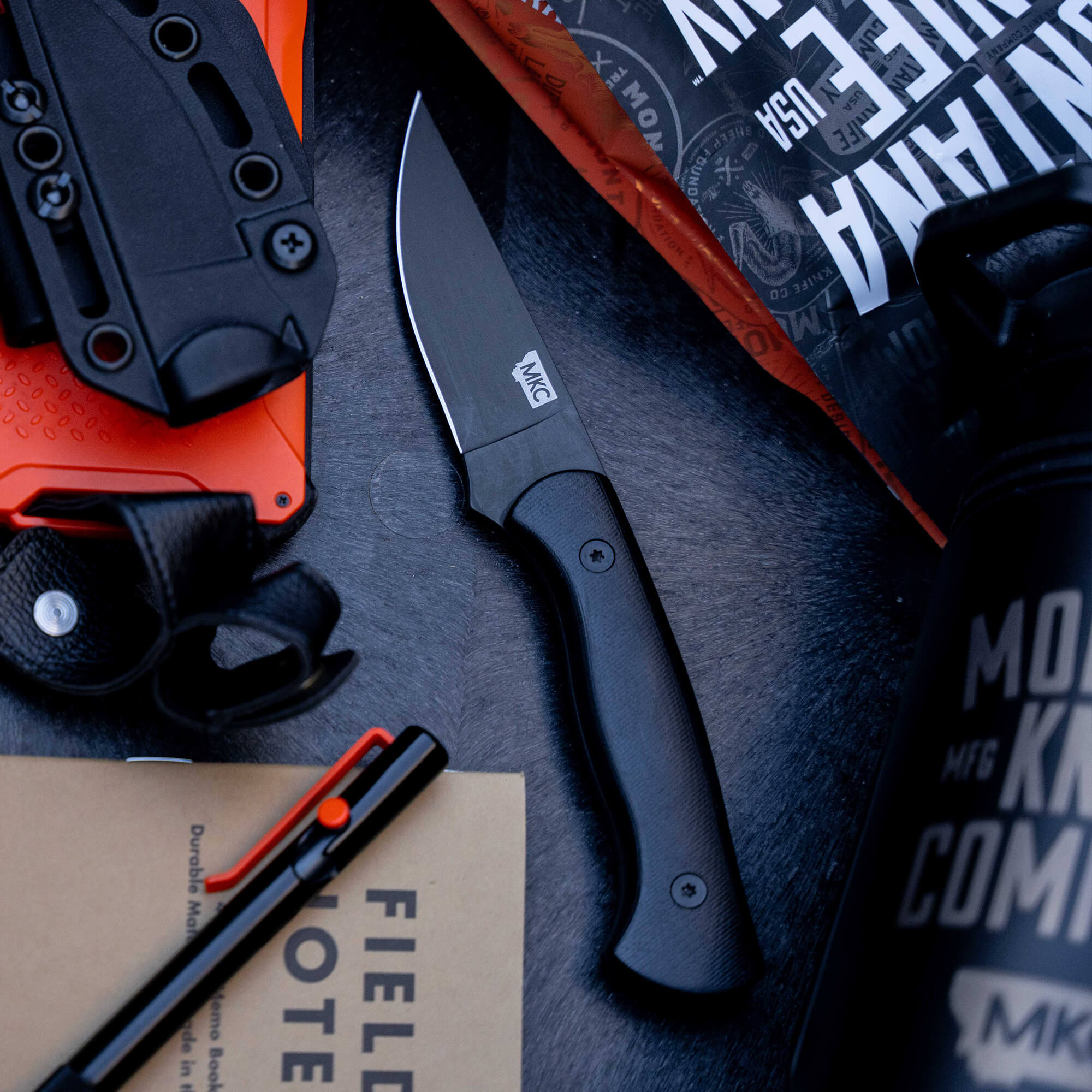 Alt text: "Blackfoot 2.0 tactical knife displayed alongside a black sheath, multitool, field notes, and a water bottle, all arranged on a dark textured surface."