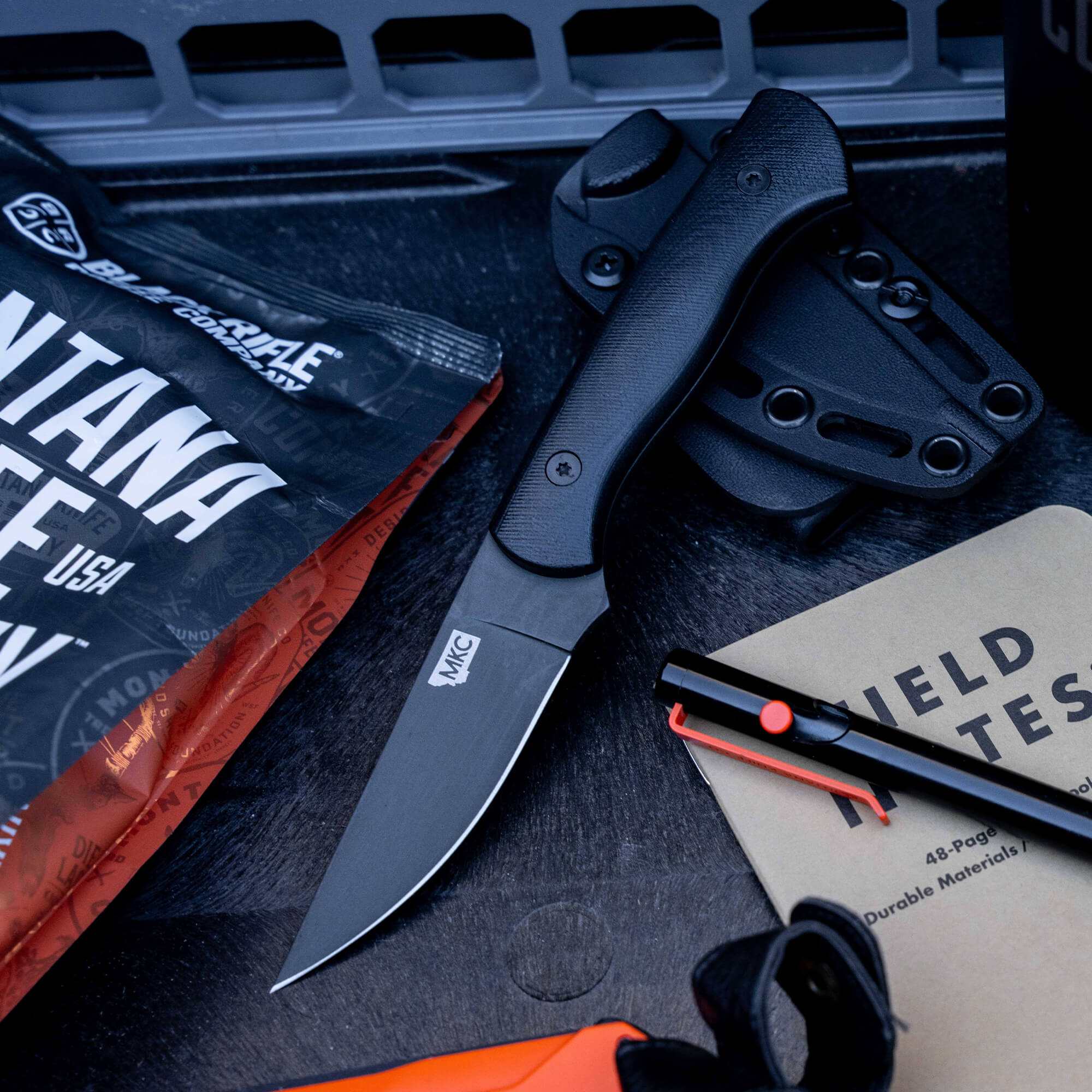 Alt text: Blackfoot 2.0 tactical knife in black, displayed with its sheath and other survival gear including a Field Notes notebook and a red fire starter, all laid out on a dark, textured surface.