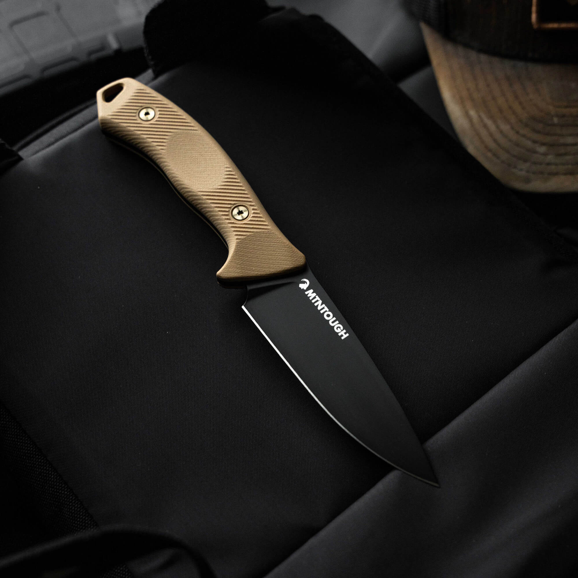 MTNTOUGH TF24 knife featuring a sleek black blade and a textured tan handle, resting on a dark surface.