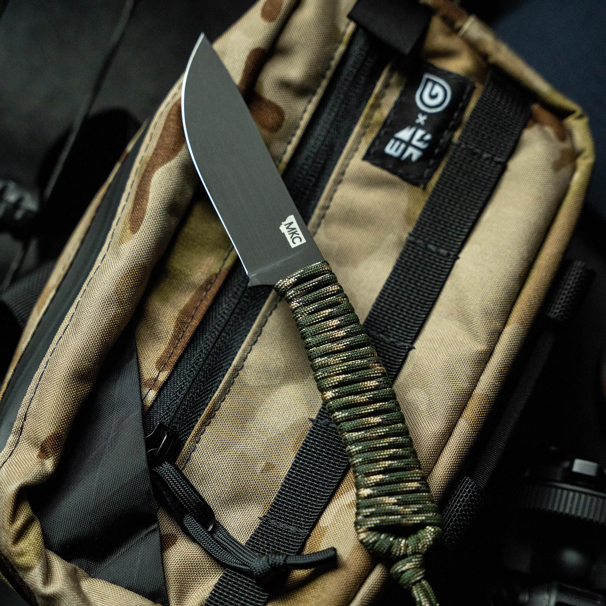 Alt text: "Close-up view of a robust tactical knife with a matte gray blade and green paracord wrapped handle, resting on a camouflage backpack with visible storage pockets and straps."