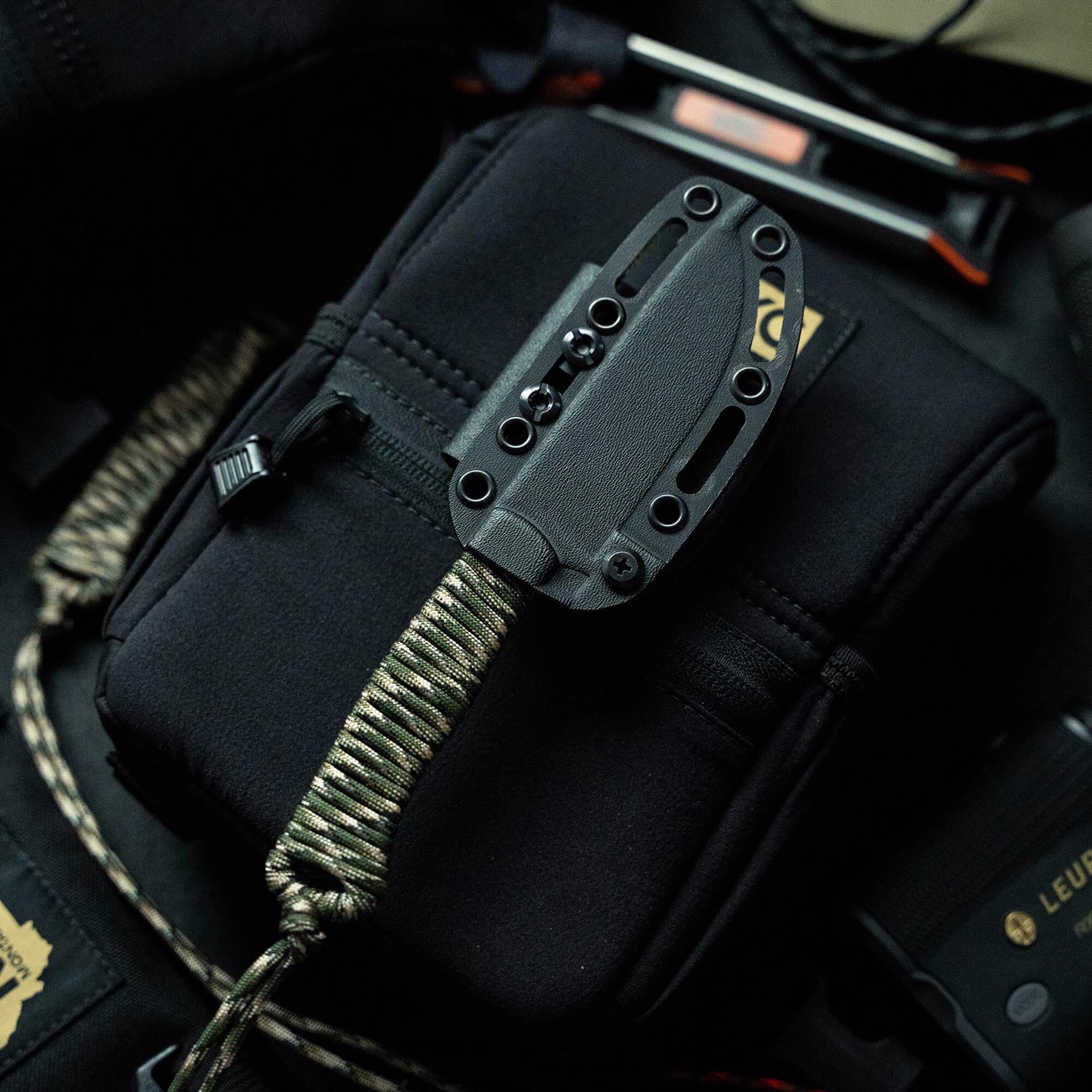 Alt text: Close-up view of a tactical knife in a black sheath attached to a cord and gear, highlighting its rugged design and the detailed workmanship of its hilt and sheath, part of the Wild Sheep Foundation Stoned Goat 2