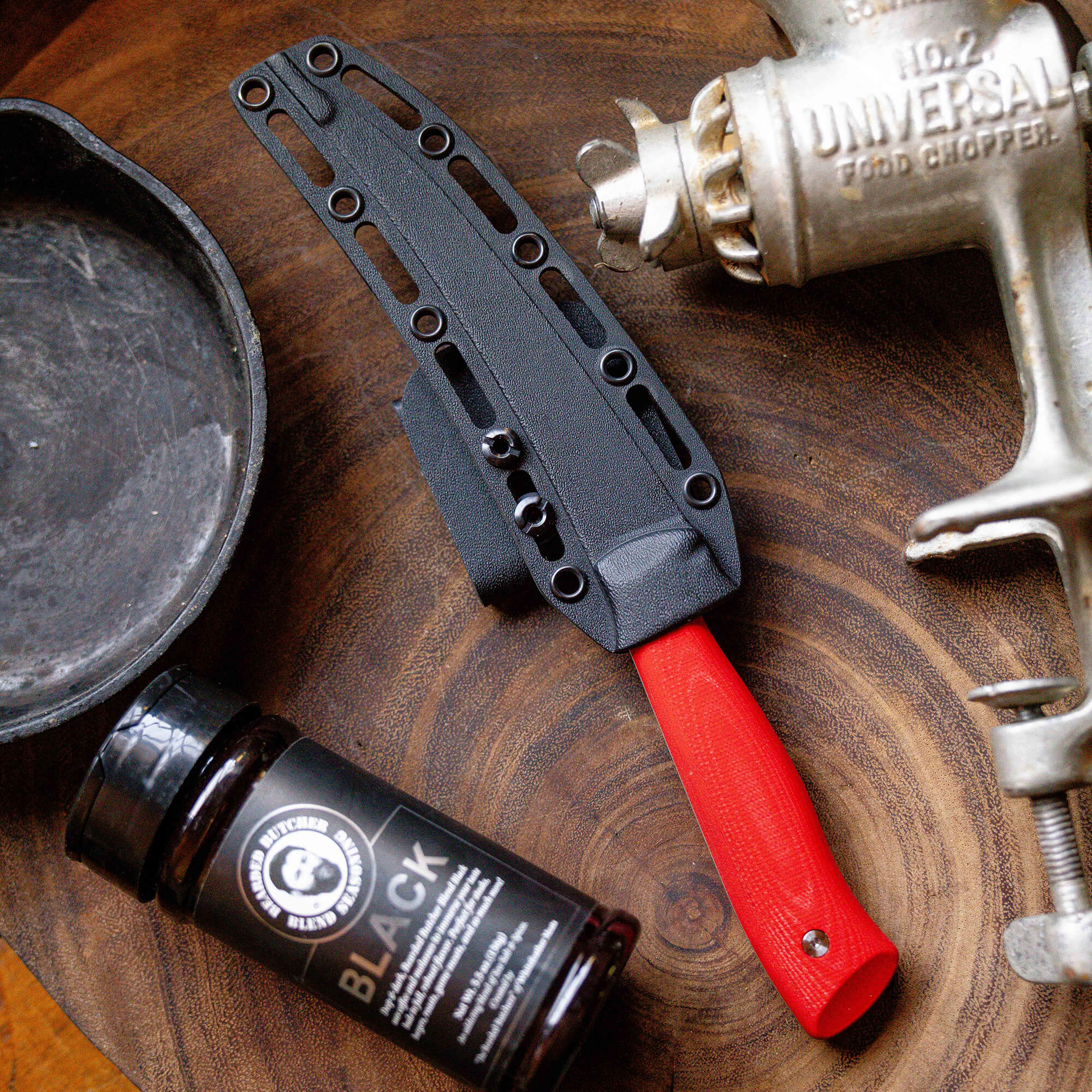 Alt text: "Bearded Butchers boning butcher knife with a red handle and black sheath, displayed next to a jar of Black seasoning blend and a vintage Universal food chopper on a wooden surface."