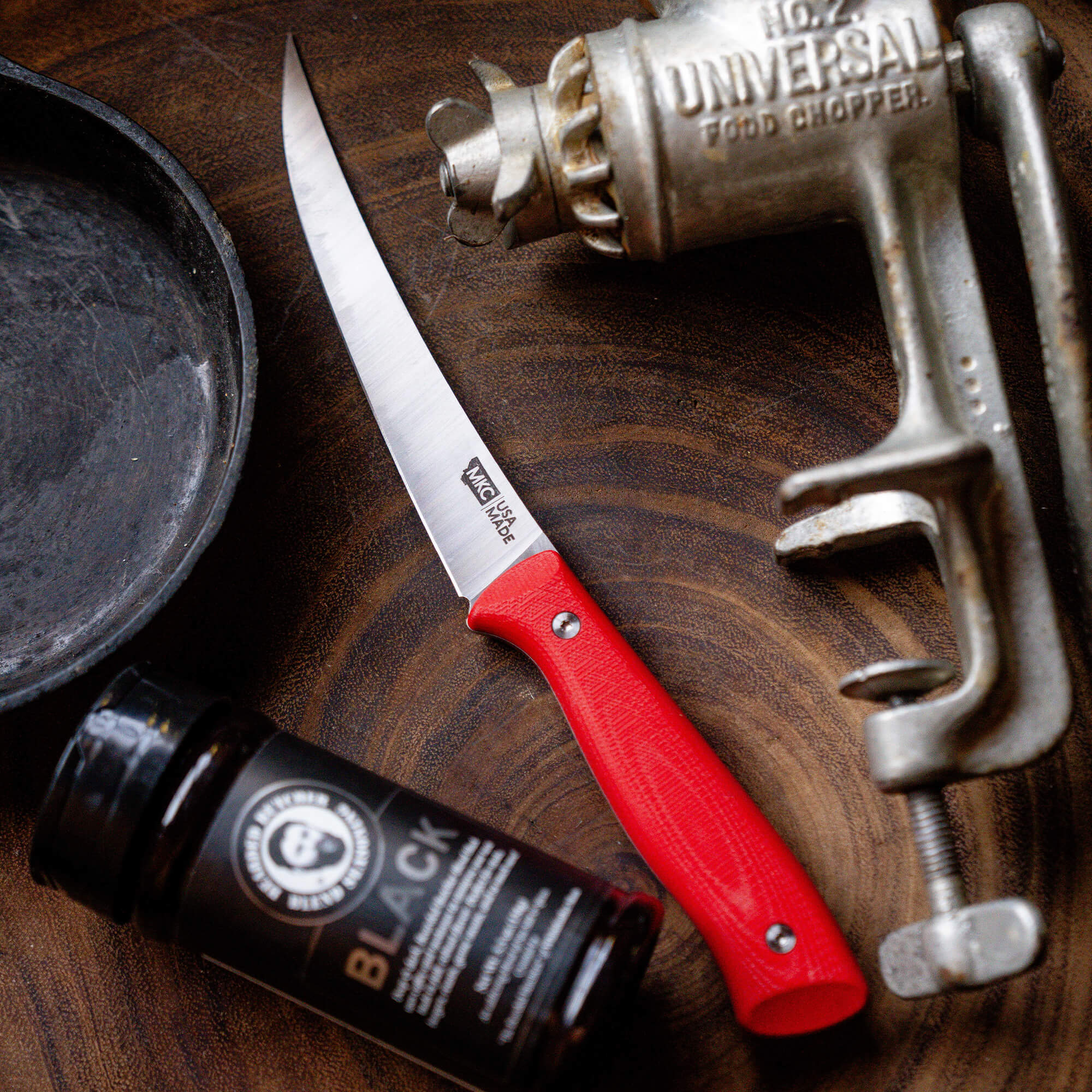 "Bearded Butchers Boning Butcher Knife with red handle, displayed on a wooden background next to a vintage metal food chopper and a black seasoning bottle."